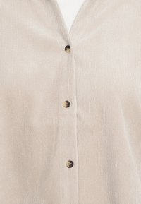 Jasny beżowy koszula z guzikami, o prążkowanej strukturze, z dekoltem w serek, z czarnymi i kremowymi guzikami zapinającymi przód.