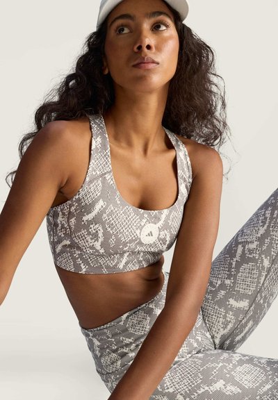 Vrouw met lang krullend haar die een grijs-witte sportbeha met slangenprint en leggings draagt, zittend en omhoog kijkend, een witte pet op haar hoofd.