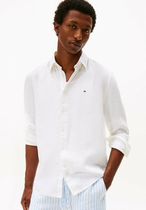 Uomo con indosso una camicia bianca a bottoni con le maniche arrotolate e pantaloni a righe azzurro chiaro, in piedi con una mano in tasca.