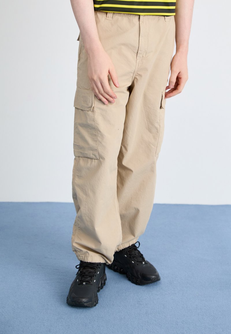 Beige cargo bukser med sidelommer, afslappet pasform og manchetter, parret med sorte træningssko. Neutral baggrund og blå gulv.