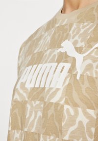 Beige kortermet skjorte med camouflage-mønster og en stor hvit "PUMA"-logo på forsiden, laget av et mykt, strukturert stoff.