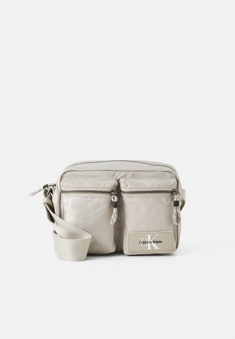 Beige nylon crossbody bag with two front zippered pockets, adjustable strap, and logo patch. Smooth texture with contrasting hardware accents.