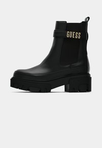 Sorte ankelstøvler laget av glatt lær med en kraftig plattformsåle. Har et fremtredende gull "GUESS"-logo og elastiske sidepaneler.