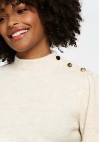 Maglione color crema con collo alto a costine e tre bottoni dorati sulla spalla. Texture morbida e design aderente.