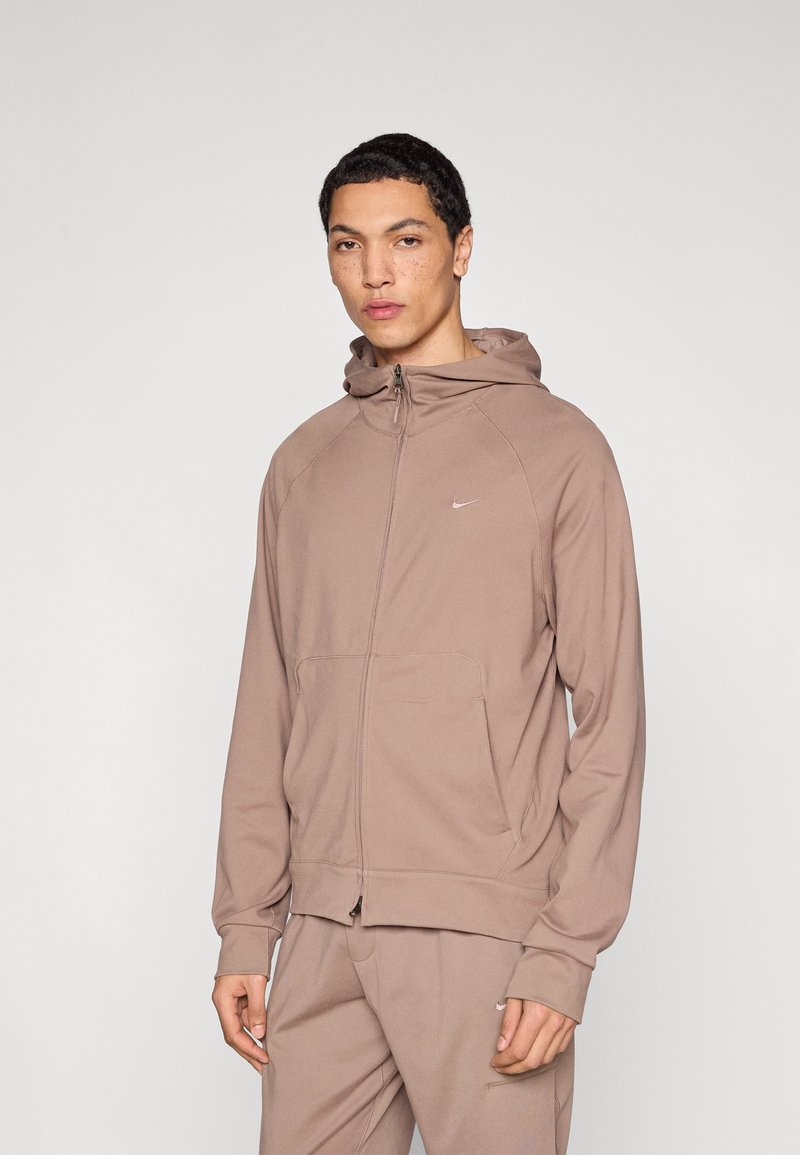 Καφέ φούτερ με φερμουάρ, κατασκευασμένο από μαλακό ύφασμα, με τσέπη καγκουρό, ελαστικά μανίκια και μικρό λογότυπο στο στήθος.