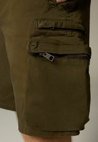 Olíva zöld cargo rövidnadrág tartós anyagból, oldalsó zsebekkel, cipzárakkal és varrott szegélyekkel. Funkcionális dizájn utilitarista részletekkel.