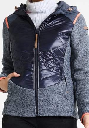 Femme portant une veste zippée avec un panneau avant matelassé bleu marine, des manches en maille grise et des détails de couleur pêche sur un pull roulé blanc.