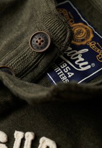 Olivengrøn sweater med en tekstureret strik, der har en mørk knap mærket "SUPERDRY" og et blåt mærke med hvid skrift og detaljer.
