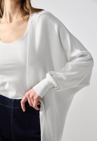 Vitt stickat cardigan med vida ärmar, bärs över en figurnära vit skjorta och ihop med mörka jeans. Slät textur, avspänd passform.