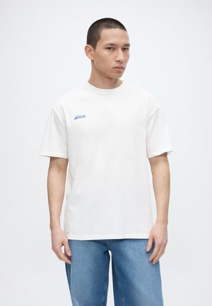 Νέος άνδρας με κοντά μαλλιά που φορά λευκό μπλουζάκι ARKK και μπλε τζιν, στέκεται μπροστά σε απλό λευκό φόντο.