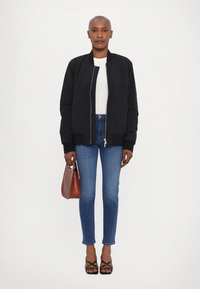 Zwarte bomberjack, ribbelmanchetten en kraag; witte gestructureerde top; blauwe skinny jeans; bruine handtas; zwarte sandalen met bandjes; neutrale achtergrond.