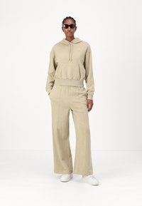 Beige hoodie med ficka framtill, parat med vida byxor. Båda tillverkade av mjukt tyg; bärs med vita sneakers och solglasögon.