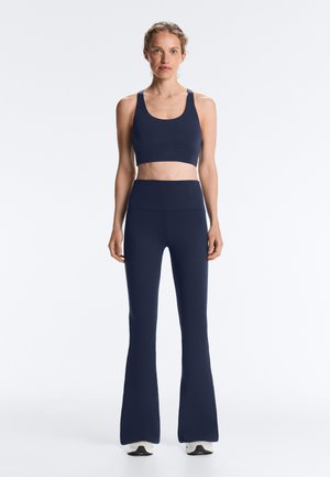 Femeie în picioare purtând un sutien sportiv bleumarin și colanți evazați cu talie înaltă, alături de adidași albi, pe fundal alb simplu.