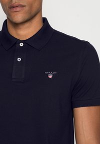 Marina blå pikétröja i bomull med klassisk krage, utrustad med två knappar och en liten GANT-logga broderad på bröstet.