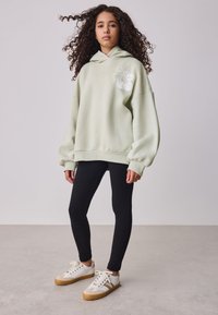 Világoszöld kapucnis pulóver elején grafikával, bő szabású, bordázott mandzsettákkal, fekete leggingszel és barna részletekkel díszített fehér tornacipőkkel párosítva.