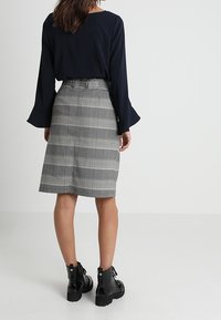 Schwarzes langärmliges Oberteil mit ausgestellten Manschetten, grauer karierter knielanger Rock und schwarze Stiefeletten mit klobigen Sohlen und Schnürungen.