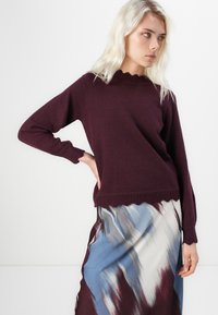 Burgundská pletená svetr s vlnitým výstřihem a rukávy, doplněný o splývavou sukni s abstraktními modrými a burgundskými vzory.