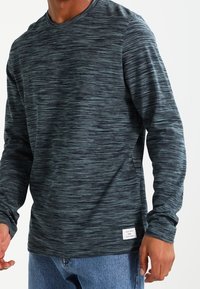 Mörkblå långärmad T-shirt med rund hals och texturerad, svepande mönster. Inkluderar en vit etikett vid fållen. Tyget verkar mjukt och bekvämt.