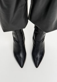 Botas de cuero negras de punta afilada hasta el tobillo, combinadas con pantalones anchos negros recortados, sobre un suelo blanco.