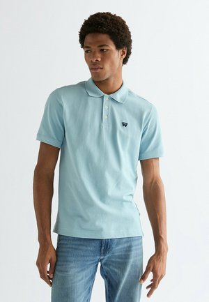 Mann trägt hellblaues Polohemd mit kurzen Ärmeln und drei Knöpfen sowie blaue Jeans, steht vor einem einfarbigen weißen Hintergrund.