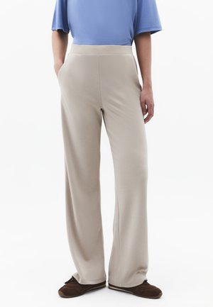 Osoba ubrana w beżowe spodnie o szerokich nogawkach, niebieską bluzkę z krótkim rękawem i brązowe buty na sznurowadła, stojąca z rękami w kieszeniach.