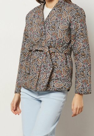 Woman wearing a belted, long-sleeve jacket with a multicolored floral and paisley pattern and light blue jeans, standing against a plain background.