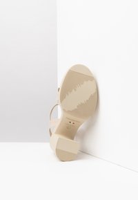 Sandalo con tacco alto beige con suola texturizzata, design aperto sul retro e sottili cinturini. Il materiale appare liscio con una finitura opaca.