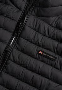 Czarna puchowa kurtka z poziomym pikowaniem, z zamkiem błyskawicznym z przodu i małą metką z logo Superdry w czerwono-białych kolorach na piersi.