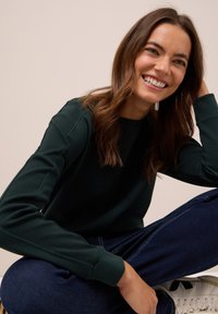 Mörkgrön sweatshirt med ribbade ärmar, rund halsringning och avslappnad passform, i kombination med mörka jeans och vita sneakers. Slät textur.