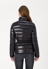 Svart, glansig vadderad jacka med horisontell quiltad design, hög krage och midjebälte. Bärs över figurnära mörka jeans.