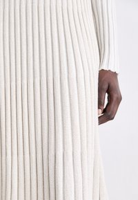 Kermanvärinen ribbed-mekko, jossa on pystysuorat poimutukset. Pehmeä rakenne, pitkät hihat ja lievä levennys helmaosassa. Korostettu käsi näkyvissä.