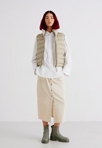 Chaleco acolchado beige, camisa blanca de botones y falda crema con botones frontales. Botines verdes hasta el tobillo. Diseño y paleta de colores limpios y minimalistas.