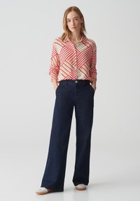 Camicia a righe rossa e crema con bottoni, abbinata a pantaloni in denim a gamba larga di colore blu scuro. Tessuto morbido con vestibilità rilassata e due tasche laterali.