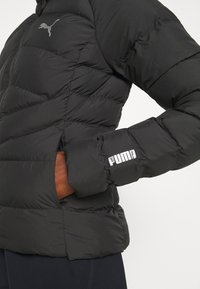 Černá péřová bunda s prošívaným designem, vybavená zipovým zapínáním a bílým logem "PUMA" na manžetě. Vyrobena z lehkého, izolačního materiálu.