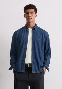 Chemise en denim bleu foncé avec un col boutonné, une poche poitrine et une coupe décontractée. Portée par-dessus un t-shirt blanc, associée à un pantalon noir.