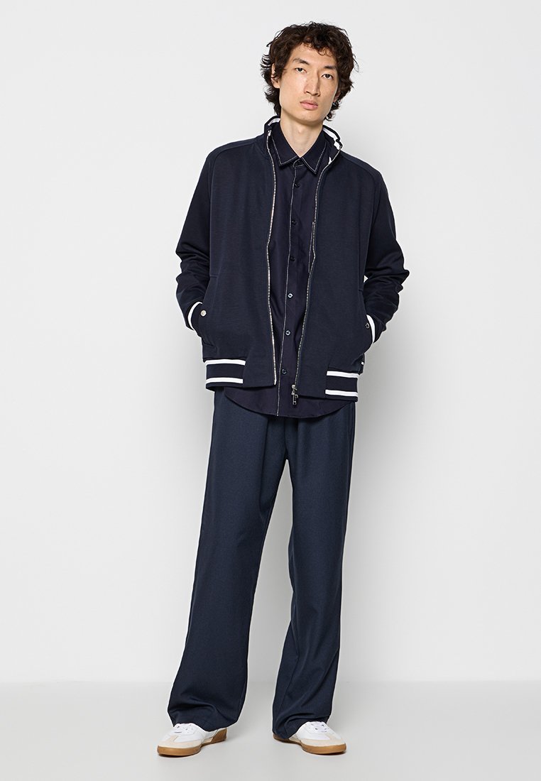 Granatowa kurtka na zamek z białymi paskami przy mankietach, zestawiona z ciemną koszulą na guziki i szerokimi spodniami. Białe buty dopełniają stylizację.