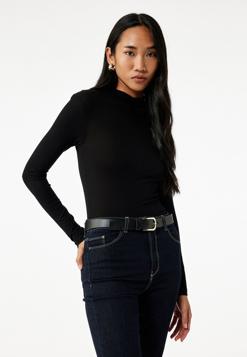 Zwarte langemouwen top met een hoge hals en gathered detail, gecombineerd met donkere denim jeans en een zwarte riem met een zilveren gesp.