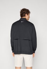 Svart hettegenser med høy krage, teksturert stoff og lagdelt bakdesign. Har liten hvit Nike-logo på øvre del av ryggen.