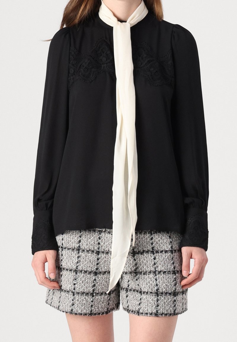 Czarna bluzka z koronkowymi detalami, wysokim kołnierzem i kokardą w kontrastowym kolorze kości słoniowej. Połączona z teksturowanymi, szarymi szortami w kratę.