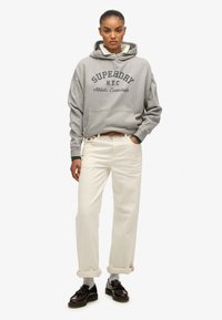 Sudadera gris con "SUPERDRY N.Y.C. Athletic Essentials" impresa en letras oscuras, combinada con pantalones blancos y zapatos negros, en una pose de pie.