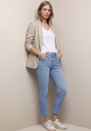 Kobieta stojąca przy beżowej ścianie, ubrana w beżową marynarkę, białą koszulkę z dekoltem w serek, jasnoniebieskie dżinsy i białe trampki, z rękami w kieszeniach.