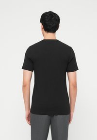 Άνδρας που φοράει απλό μαύρο κοντομάνικο μπλουζάκι και γκρι παντελόνι, στέκεται με την πλάτη προς το μέρος του θεατή, μπροστά σε λευκό φόντο.