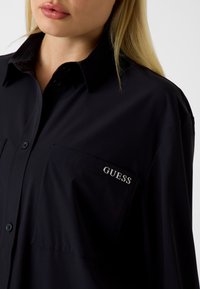 Zwart shirt met lange mouwen en een button-down kraag, voorzien van een enkele zak en een zilveren "GUESS"-logo op de borst. Gladde stoftextuur.