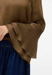 Brun, strukturerad blus med utsvängda ärmar. Nära bild visar vågiga kanter och ett mjukt tyg. Bärs med en denimkjol.