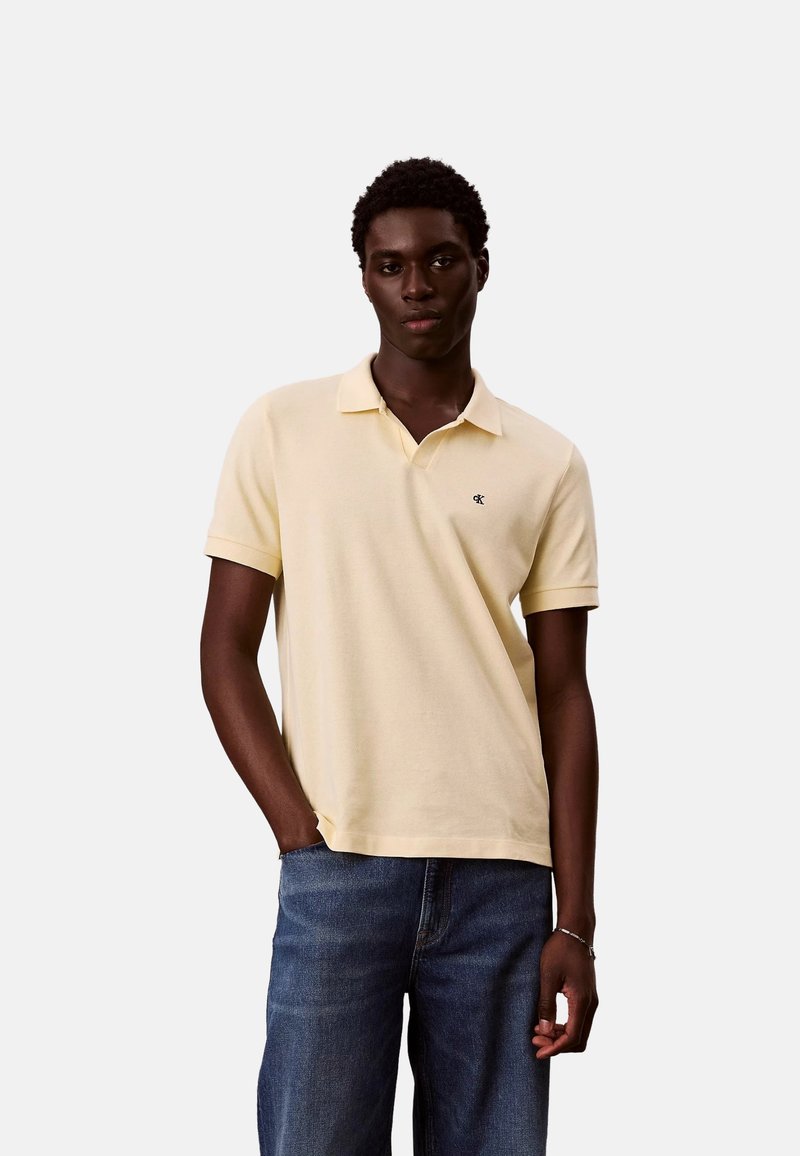 Jeune homme debout, une main dans la poche, portant un polo à manches courtes jaune pâle et un jean bleu, sur fond uni.