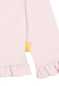 Helles rosa Langarmoberteil mit Rüschensäumen, aus weichem Stoff gefertigt. Es besitzt ein kleines gelbes Etikett mit Markenaufschrift an der Seite.