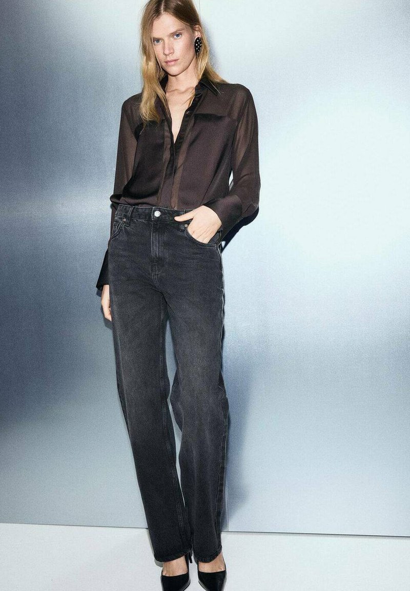 Black sheer button-up shirt paired with high-waisted dark gray straight-leg jeans. Model wearing black heels and oversized black earrings.