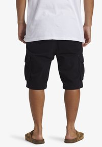 Zwarte cargoshorts met zijzakken, knielange snit en een relaxed fit, gecombineerd met bruine slidesandalen en een wit T-shirt.