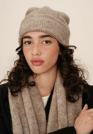 Bonnet en tricot côtelé beige et écharpe texturée assortie, tous deux fabriqués dans un matériau doux, portés par un modèle avec des cheveux bouclés.