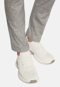 Vita idrottsskor med texturerade ovandelar och snören, kombinerat med grå, något avsmalnande byxor med en subtil melerat mönster.